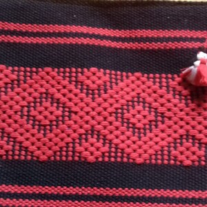 Puede incluir: Una tela tejida negra y roja con una borla blanca. La tela tiene un patrón geométrico y está hecha con un hilo grueso.