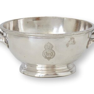 May include: A silver bowl with two handles and a decorative emblem. The bowl has a wide, shallow design with a rounded base and a polished finish. The emblem appears to be a crest or coat of arms.