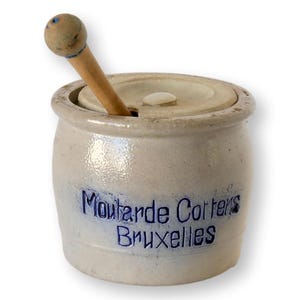 May include: A vintage stoneware mustard pot with a lid and wooden spoon. The pot is off-white with blue lettering that reads "Moutarde Cotters Bruxelles". The wooden spoon has a rounded end.