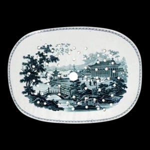 May include: Oval, white ceramic plate with a blue and white Asian landscape design. The plate features a series of small holes and a decorative border. The landscape includes pagodas, bridges, and boats, creating a serene scene.
