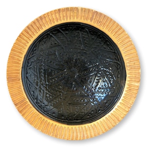 May include: A round decorative plate with a textured, light brown rim and a black center. The center features an embossed geometric design. The plate is set against a white background.
