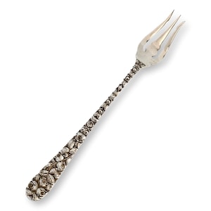 May include: A silver-toned metal fork featuring a three-pronged head and a handle intricately embossed with a detailed floral pattern of roses and leaves. The metallic surface has a polished finish with dark recessed areas highlighting the design.