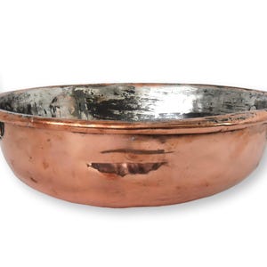 May include: A copper bowl with two arched handles. The bowl has a rounded shape with a wide opening and a slightly textured surface. The interior has a silver-colored finish, and the exterior is a warm copper color.