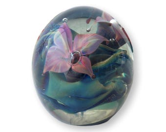 May include: A clear glass paperweight with a floral design. The paperweight has a rounded, oval shape. Inside, a pink and purple flower is suspended within swirling blue and green glass. Small air bubbles are visible.