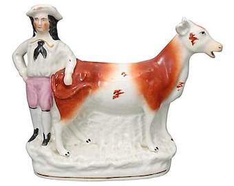 May include: A vintage ceramic figurine depicting a figure in a white shirt, pink shorts, and a hat, standing next to a cow. The cow is white with red-brown patches. The figurine is on a white base.