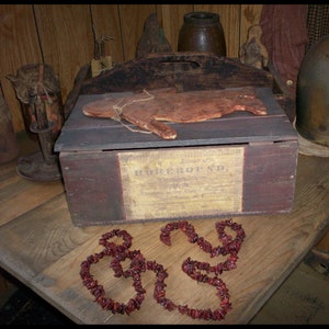 May include: A wooden box with a label that reads "HOREBOUND" and a red, white, and brown color scheme. The box is sitting on a wooden table with a red cranberry garland in the shape of a heart.