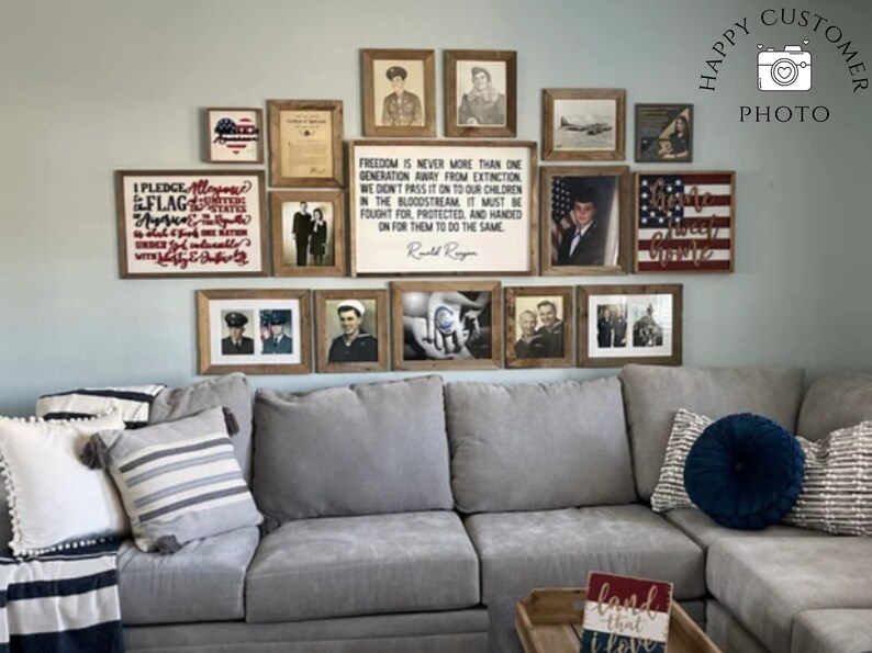 May include: A gray sectional sofa with pillows and a throw blanket. The wall behind the sofa is decorated with framed pictures and signs with patriotic themes, including the American flag, a quote about freedom, and a sign that says "Home Sweet Home".
