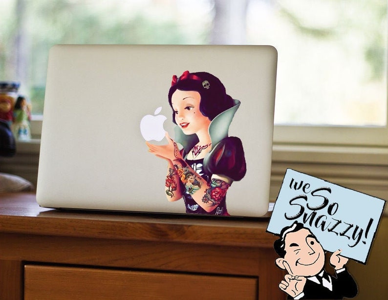 May include: A laptop with a sticker featuring a Snow White-inspired character with tattoos, holding an Apple logo. The character has a red bow and a white collar. A sign with the text "we So Snazzy!" is in the corner.