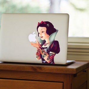 May include: A laptop with a sticker featuring a Snow White-inspired character with tattoos, holding an Apple logo. The character has dark hair, a red bow, and a black and white outfit. The laptop is on a wooden desk.
