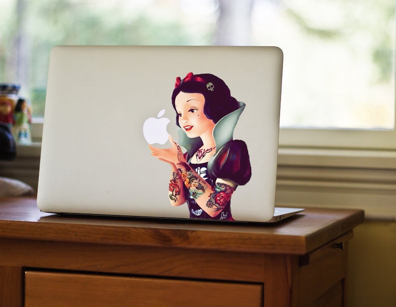 May include: A laptop with a sticker featuring a Snow White-inspired character with tattoos, holding an Apple logo. The character has dark hair, a red bow, and a black and white outfit. The laptop is on a wooden desk.