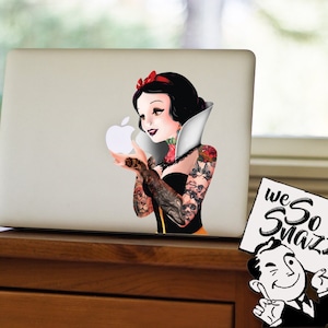 May include: A white laptop with a sticker of Snow White with tattoos holding a white apple. The sticker is on the top half of the laptop. The laptop is sitting on a wooden surface. A white sign with the text "we So Snazzy!" is on the right side of the image.