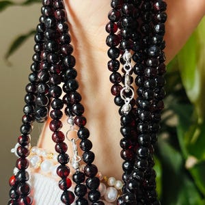 May include: Several strands of dark red and black beaded necklaces with silver clasps. The beads are round and appear to be made of a polished stone. A white sweater is visible in the background.