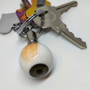 May include: A silver key chain with a realistic eyeball charm. The eyeball is white with a brown iris and a light brown sclera. The keychain also includes a silver key and a guitar-shaped key charm.