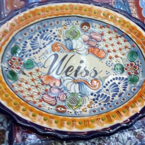 Könnte beinhalten: Ovale Keramikplatte mit handgemaltem Blumendesign in Blau, Orange und Grün. Die Platte hat einen gewellten Rand und der Name "Weiss" ist in Schreibschrift in der Mitte geschrieben.