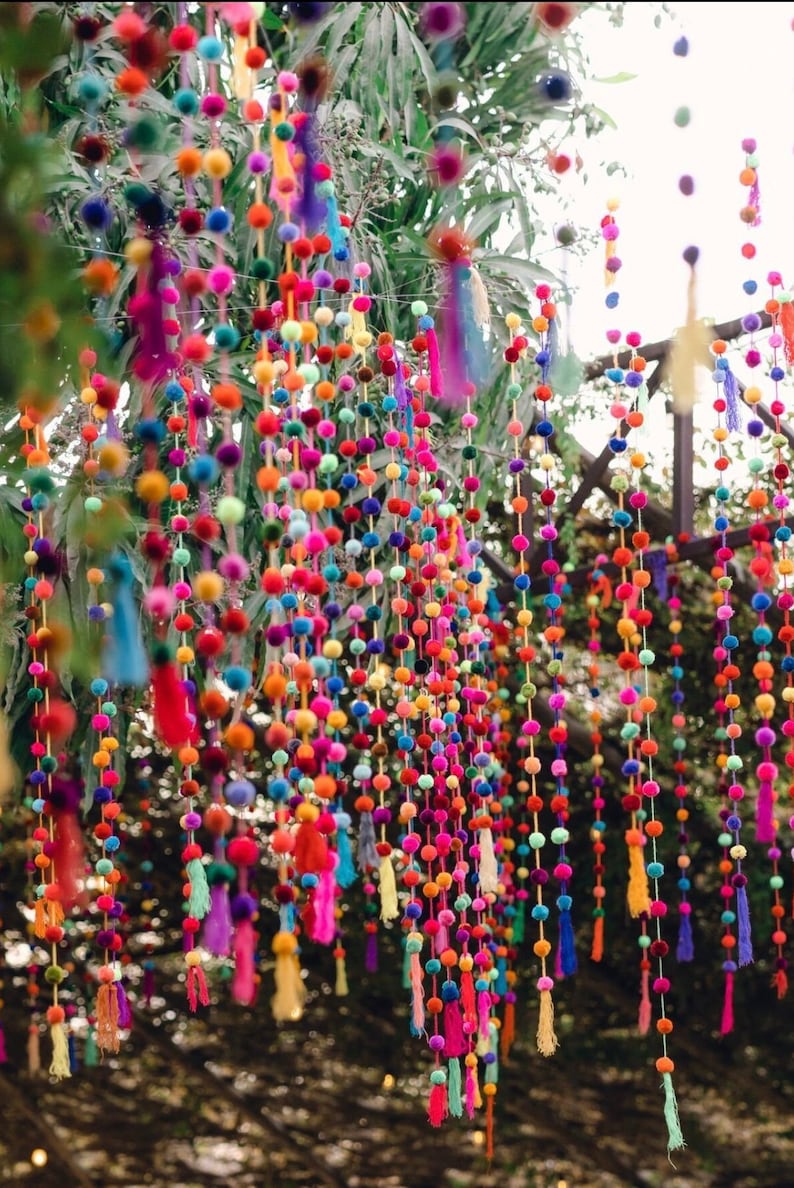 May include: A colourful garland of pom-poms and tassels hangs from a tree branch. The pom-poms are in various shades of pink, purple, blue, green, yellow, and orange. The tassels are in matching colours and add a whimsical touch to the garland.