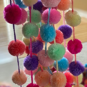 May include: A colorful garland of fluffy pom-poms in various shades of pink, blue, green, and purple. The pom-poms are strung together on white yarn.