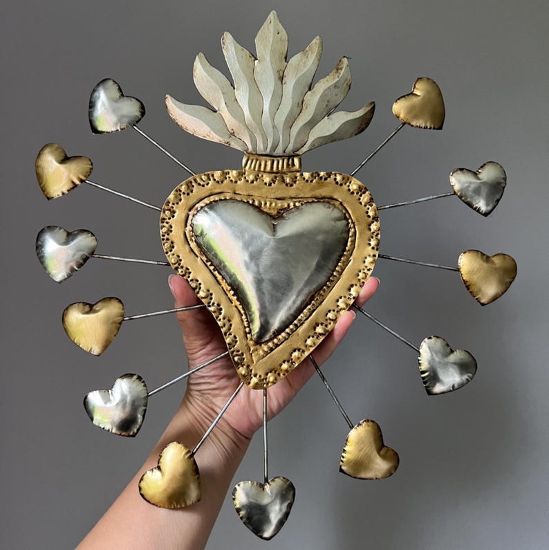 May include: A gold and silver metal heart with a white flame on top and 12 smaller hearts surrounding it. The hearts are attached to the larger heart with thin wires.