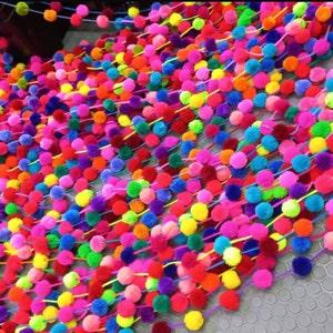 May include: A colourful garland of pom-poms in various shades of pink, purple, blue, green, yellow, orange, and red. The pom-poms are strung together on a thin string.