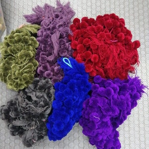 May include: Assortment of colourful pom-pom trim in various colours including green, purple, red, blue, and black. The trim is made of soft, fluffy yarn and is perfect for adding a touch of whimsy to any project.