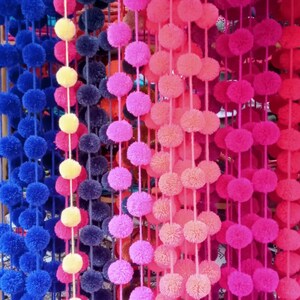 May include: A close-up of a wall decorated with colourful pom-pom garlands. The garlands are made of fluffy yarn and are strung together in a variety of colours, including blue, yellow, pink, and red.