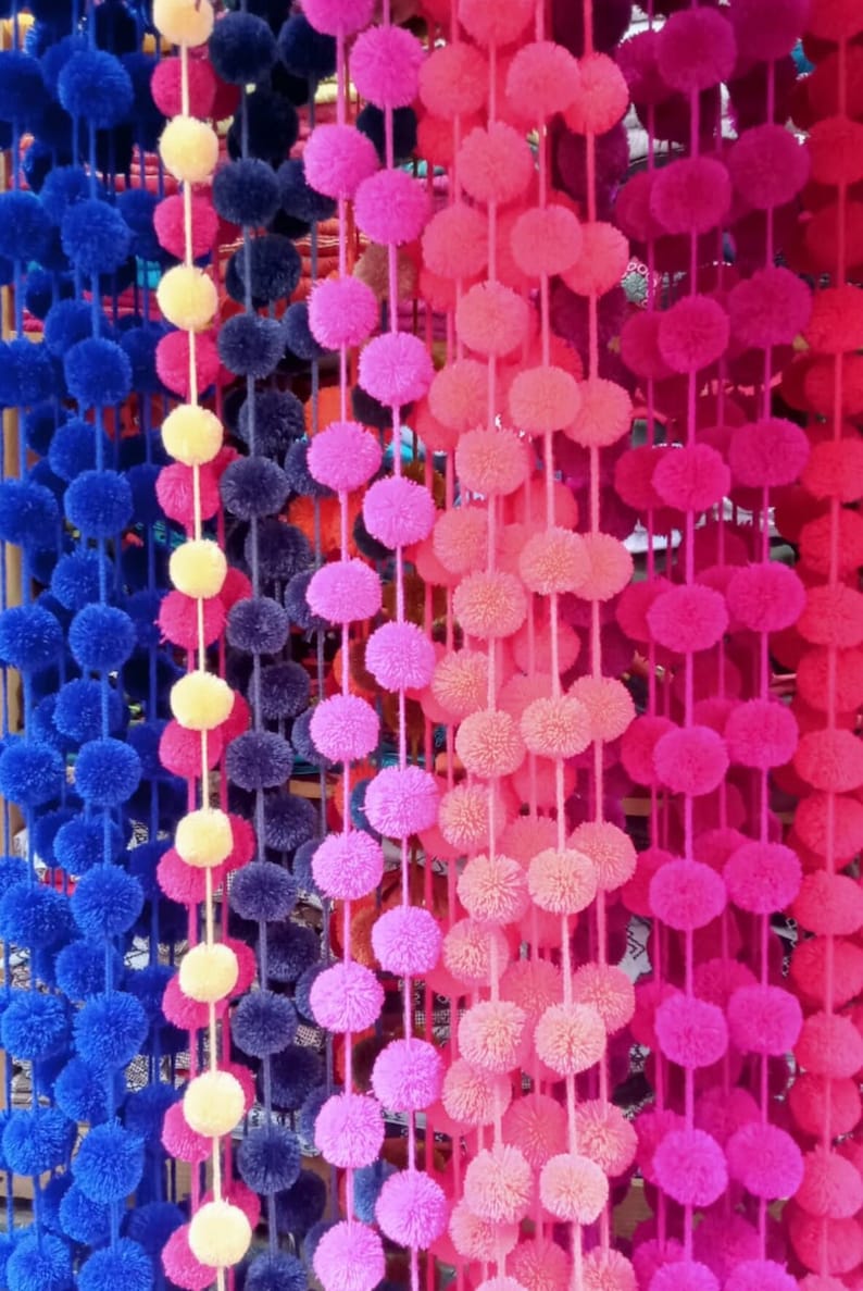 May include: A close-up of a wall decorated with colourful pom-pom garlands. The garlands are made of fluffy yarn and are strung together in a variety of colours, including blue, yellow, pink, and red.