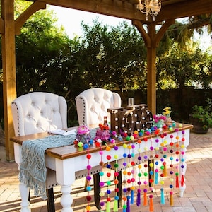 May include: A wedding sweetheart table decorated with a white tablecloth, a blue crocheted runner, and a garland of colourful pom-poms with tassels. The table is set with two white chairs and a wooden sign that reads "LOVE".