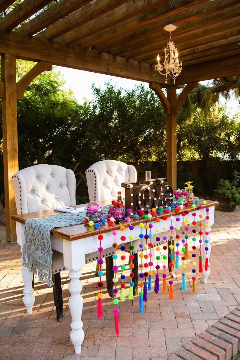 May include: A wedding sweetheart table decorated with a white tablecloth, a blue crocheted runner, and a garland of colourful pom-poms with tassels. The table is set with two white chairs and a wooden sign that reads "LOVE".