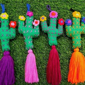 Può includere: Quattro ornamenti a forma di cactus in feltro fatti a mano con nappe colorate. Ogni cactus è verde con dettagli ricamati e sormontato da un fiore vibrante. Le nappe sono viola, rosa, bordeaux e arancioni. Gli ornamenti sono esposti su una superficie erbosa verde.