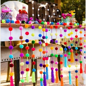May include: A table decorated with colourful pom pom garlands with tassels. The garlands are strung across the table and hang down to the floor. The table is white and has a wooden top. The garlands are made of yarn and are in a variety of colours, including red, orange, yellow, green, blue, purple, and pink. The word "LOVE" is spelled out in large, brown letters with white lights behind them. The letters are sitting on the table in front of the garlands.