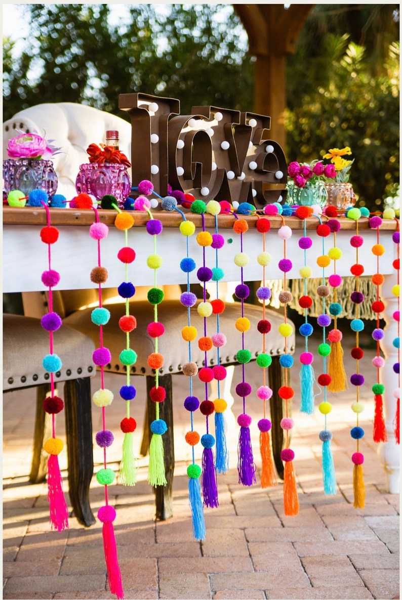May include: A table decorated with colourful pom pom garlands with tassels. The garlands are strung across the table and hang down to the floor. The table is white and has a wooden top. The garlands are made of yarn and are in a variety of colours, including red, orange, yellow, green, blue, purple, and pink. The word "LOVE" is spelled out in large, brown letters with white lights behind them. The letters are sitting on the table in front of the garlands.