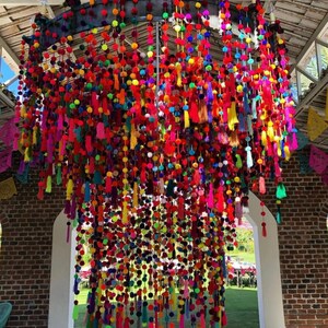 May include: A large, colourful chandelier made of pom-poms and tassels. The chandelier is hanging from the ceiling of a room with brick walls and a white archway.