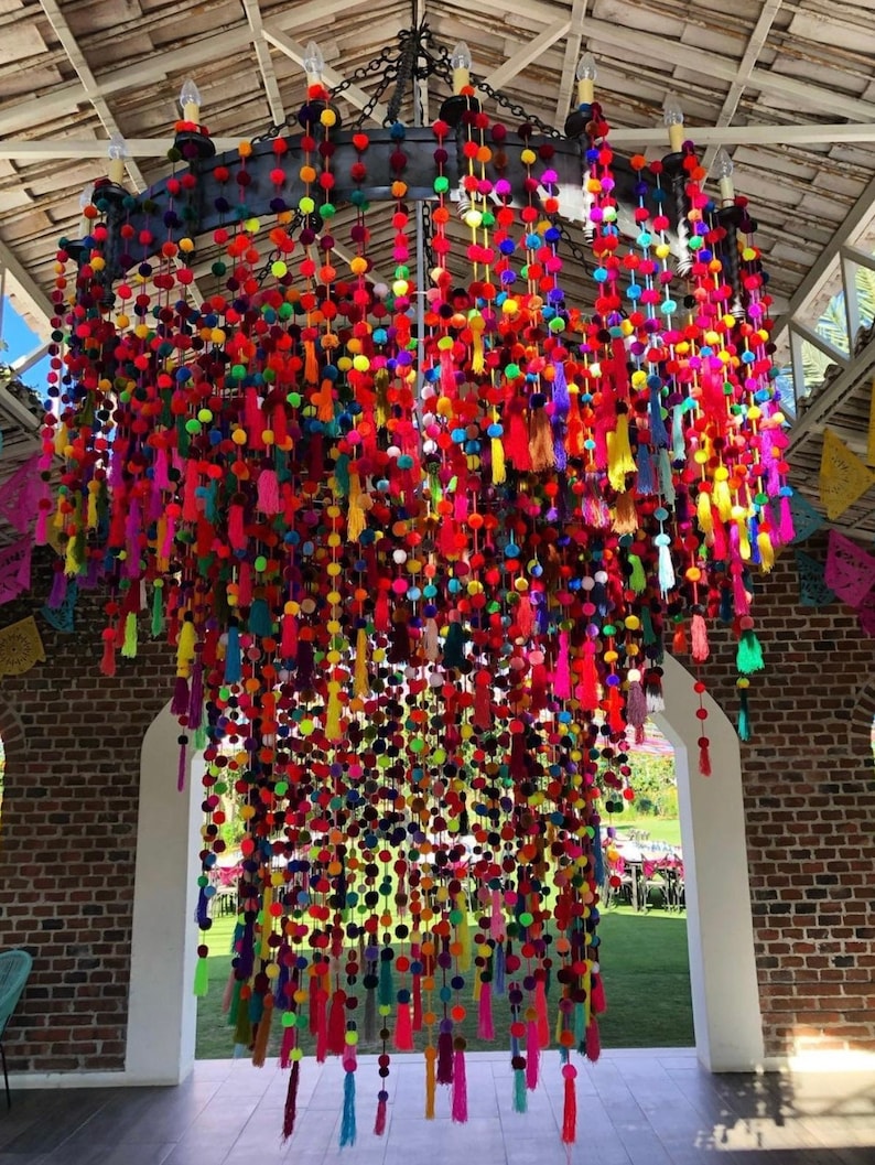 May include: A large, colourful chandelier made of pom-poms and tassels. The chandelier is hanging from the ceiling of a room with brick walls and a white archway.