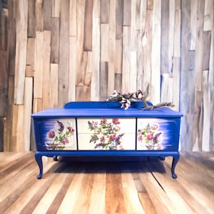 Op de afbeelding: Een rechthoekige blauwe houten kist met vier cabriolepoten, voorzien van twee zijkastjes en twee centrale lades. De voorpanelen zijn versierd met bloemmotieven in roze, paarse en groene tinten. Een takje met bloemen ligt bovenop de kist.