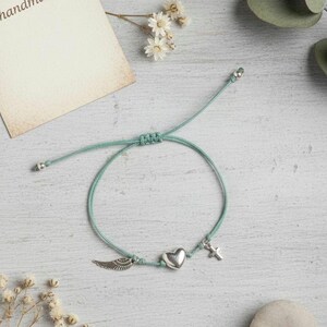 Peut inclure: Bracelet réglable avec un cordon vert d'eau. Il comporte des breloques argentées : une aile, un cœur et une croix. Le mot "handmade" est visible sur une petite carte. Bijou délicat.