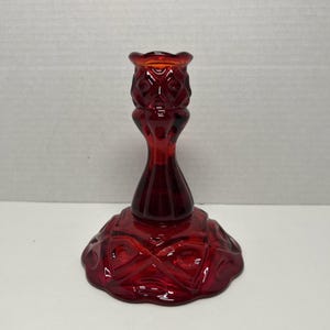 Peut inclure: Un bougeoir en verre rouge profond présentant un motif géométrique à facettes sur la large base circulaire et un design texturé assorti sur le col supérieur. La pièce possède une finition lisse et translucide ainsi qu'une ouverture évasée.