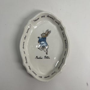 May include: An oval white ceramic trinket dish featuring a central illustration of Peter Rabbit wearing a blue jacket. The text 'Now my dears,' said old Mrs. Rabbit one morning, 'you may go into the fields or down the lane...' and 'Beatrix Potter' are printed in black along the scalloped rim.