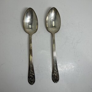 May include: Two silver-toned metal spoons featuring an elongated oval bowl and a slender handle. The handle is decorated with a detailed embossed floral pattern at the base, transitioning into a smooth, plain stem that connects to the spoon bowl.