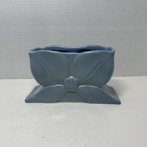 Peut inclure: Un cache-pot en céramique bleu cendré présentant un motif floral moulé avec quatre grands pétales et un bouton central. Le récipient possède une ouverture rectangulaire sur le dessus et une base large et plate, avec des détails nervurés.