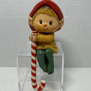 May include: A figurine of a small elf with blonde hair wearing a red hat, a yellow tunic, and green pants. The elf is holding a long, vertical candy cane with red and white diagonal stripes. The figure has pointed ears and a small orange nose.