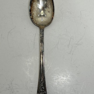 May include: A silver-toned serving spoon featuring a bowl with a decorative cutout pattern in the center. The handle is adorned with an intricate floral relief design at the base. The metal surface shows signs of natural oxidation and patina.