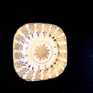 Op de afbeelding: Een wit vierkant bord met een gouden bloemmotief rond de rand en een kleiner gouden bloemmotief in het midden.