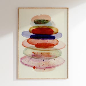 Puede incluir: Impresión artística abstracta en acuarela que presenta una pila de formas ovaladas coloridas en tonos de rosa, rojo, azul y verde. La obra de arte está enmarcada en un marco de madera clara, creando una estética moderna y minimalista.