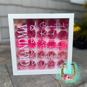 Puede incluir: Una caja de sombra con marco blanco y flores de papel rosas. Las palabras "GRANDMA" y "Beautiful, Kind, Loving, Patient, Selfless, Wise" están escritas en blanco. Las flores están dispuestas en filas.