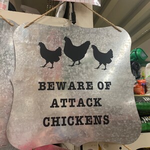 Puede incluir: Un letrero de metal plateado con el texto "Beware of Attack Chickens" y tres siluetas negras de pollos. El letrero tiene un aspecto desgastado y está colgado de una cuerda.
