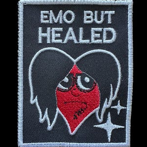 Può includere: Una toppa nera con testo bianco che recita "EMO BUT HEALED". La toppa presenta un cuore rosso con un volto, ali bianche e due stelle bianche. La toppa ha un bordo bianco.