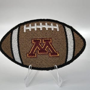 May include: An embroidered patch shaped like an American football in brown with white laces and stripes. The center features a maroon and gold block letter M logo. The patch has a thick black stitched border and is displayed on a clear stand.