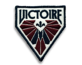 Pode incluir: Patch bordado com a palavra "VICTOIRE" em branco, sobre um desenho de formas bordô e azul marinho. O patch tem um fundo branco e uma borda azul marinho. O design é um emblema estilizado.