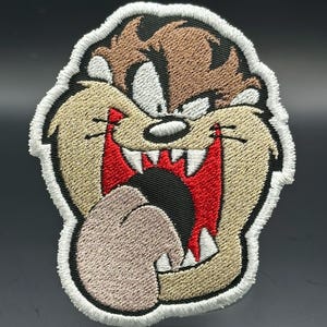 Może przedstawiać: Haftowana naszywka z postacią z kreskówki Diabła Tasmańskiego. Naszywka ma beżową twarz, brązowe włosy, czerwone usta i długi, różowy język. Projekt jest obrysowany w kolorze czarno-białym.