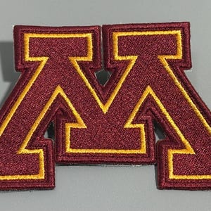 Puede incluir: Parche bordado con la letra "M" en color granate con ribete dorado. El parche tiene un aspecto texturizado y probablemente está diseñado para ser cosido o planchado sobre tela. El diseño es una letra audaz y estilizada.