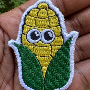Puede incluir: Parche bordado con una mazorca de maíz de dibujos animados con grandes ojos saltones. El maíz es amarillo con hojas verdes y un borde blanco. Este parche está diseñado para planchar o coser sobre tela.